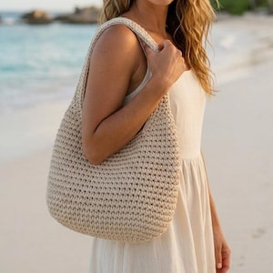 May include: A beige, crocheted shoulder bag with a rounded shape and a wide strap. The bag is made of a thick, textured yarn and is being held by a person wearing a cream-colored dress. The background is a beach scene with the ocean.
