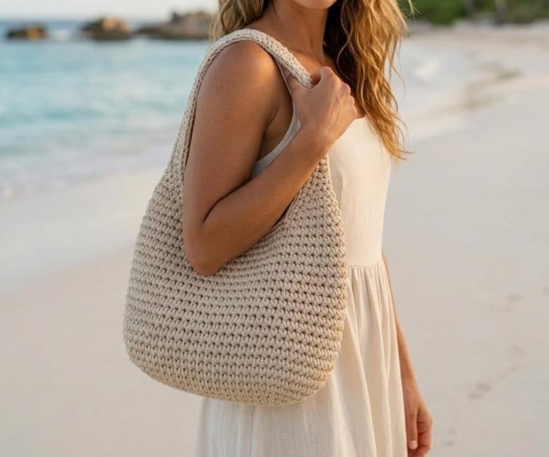 May include: A beige, crocheted shoulder bag with a rounded shape and a wide strap. The bag is made of a thick, textured yarn and is being held by a person wearing a cream-colored dress. The background is a beach scene with the ocean.