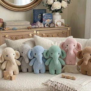 Könnte beinhalten: Eine Sammlung handgefertigter gehäkelter Elefanten in verschiedenen Pastellfarben, darunter Creme, Grau, Blau, Grün, Rosa und Braun. Die Stofftiere sind auf einem weißen Sofa mit dekorativen Kissen angeordnet und schaffen eine gemütliche Szene.