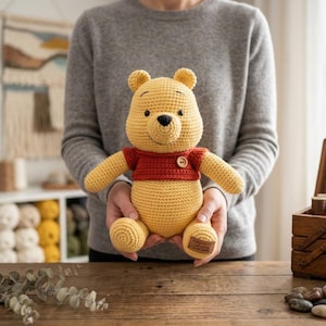 May include: A yellow crocheted bear toy wearing a red shirt with a small wooden button detail. The bear has black embroidered eyes and a black nose. A small rectangular fabric tag is attached to the bottom of the bear's left foot.