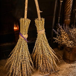 May include: Two decorative brooms crafted from dried lavender stems and natural wooden branches. Each broom features a bundle of light brown and purple lavender tied with twine, with one broom accented by a thin purple cord around the handle base.