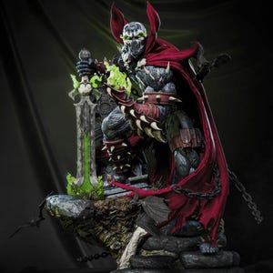 Op de afbeelding: Een gedetailleerd actiefiguur van Spawn, een stripboekpersonage, staat op een stenen voetstuk. Het figuur is gekleed in donkere bepantsering, een rode cape en houdt een groen oplichtend zwaard vast. Het figuur heeft een schedelachtig masker en is omringd door kettingen.
