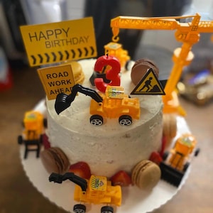May include: A round birthday cake decorated with toy construction vehicles, a number 3 candle, and the words "Happy Birthday" and "Road Work Ahead" on yellow signs. The cake is also decorated with strawberries and macarons.