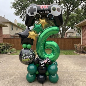 May include: A balloon arrangement featuring a large green number six, a black video game controller balloon with the text GAME ON! IT'S YOUR BIRTHDAY, a round balloon with the text NEXT LEVEL BIRTHDAY, and several gold, green, and black star balloons.