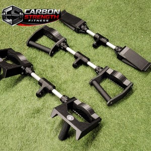 May include: Four black and silver fitness tools are arranged on a green surface. Each tool has a handle, a silver bar, and a black attachment. The Carbon Strength Fitness logo is in the upper left corner.