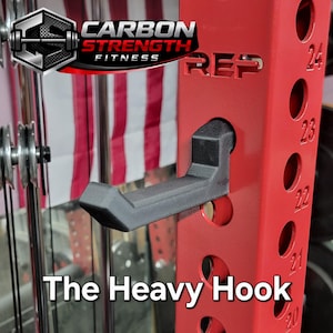 May include: A red weightlifting power rack with a black hook. The rack has the text "REP" and numbers 20-24. The text "The Heavy Hook" is at the bottom. The Carbon Strength Fitness logo is in the upper left.