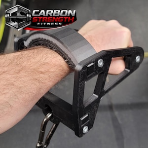 May include: Black weightlifting wrist support with the text "CARBON STRENGTH FITNESS" in red and white. The wrist support is attached to a black metal frame with a carabiner clip. The wrist support is designed to provide support during weightlifting exercises.