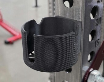 May include: A matte black cylindrical cup holder with a textured exterior surface and a U-shaped cutout on the front rim, securely attached to a vertical steel gym rack post featuring numbered circular adjustment holes along its side.