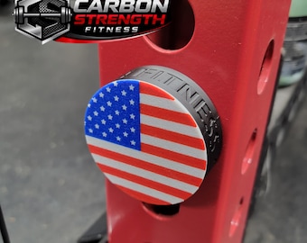 May include: A red weight rack with a round black bumper plate featuring the American flag. The bumper plate is inserted into the rack. The logo "CARBON STRENGTH FITNESS" is in the upper left corner.