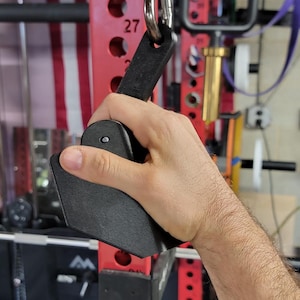 Könnte beinhalten: Schwarzer Gewichthebergriff an einem Metallring befestigt. Der Griff wird in einer Hand gehalten, mit einer strukturierten Oberfläche. Der Hintergrund umfasst ein rotes Gewichtsgestell und eine amerikanische Flagge.