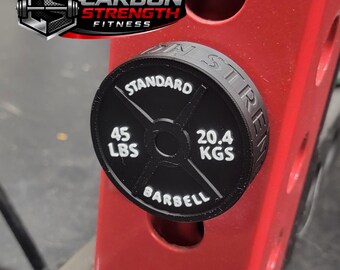 May include: Black weight plate with white text reading "STANDARD", "45 LBS", and "BARBELL". The plate also displays "20.4 KGS". The plate is attached to a red metal structure. The logo "CARBON STRENGTH FITNESS" is in the upper left corner.