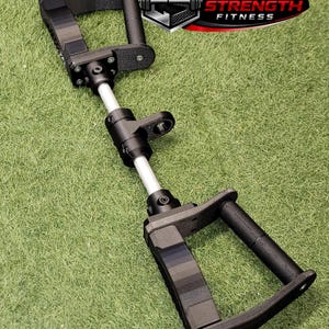 Pode incluir: Um acessório de fitness preto e prateado com duas pegas texturizadas ligadas por uma haste metálica central com um ponto de fixação. O dispositivo inclui uma placa com o logótipo e o texto CARBON STRENGTH FITNESS em letras pretas e vermelhas.