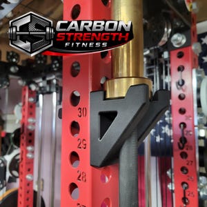 May include: A red weightlifting power rack with a black bar holder and a gold-colored bar. The rack has numbered holes for adjusting the bar height. The logo "CARBON STRENGTH FITNESS" is visible.