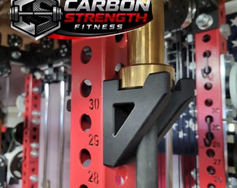 May include: A red weightlifting power rack with a black bar holder and a gold-colored bar. The rack has numbered holes for adjusting the bar height. The logo "CARBON STRENGTH FITNESS" is visible.