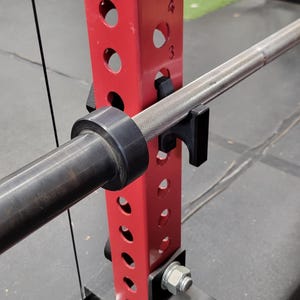 May include: A red vertical steel power rack upright with numbered circular holes, featuring a black J-hook attachment holding a metal barbell. A black barbell collar is secured on the bar, and a black pulley system component is attached at the base.