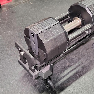 May include: Adjustable dumbbell set with a black and silver finish. The dumbbell features a textured, knurled handle and a weight adjustment mechanism. The weight plates are black and rectangular. The set includes a stand with wheels for easy movement.