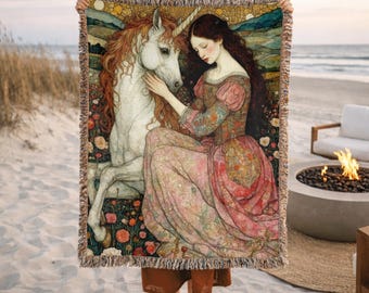May include: Woven tapestry blanket featuring a fantasy scene with a woman and a unicorn. The artwork depicts a woman in a pink dress with a unicorn. The blanket has a fringed edge and is held up against a beach backdrop.