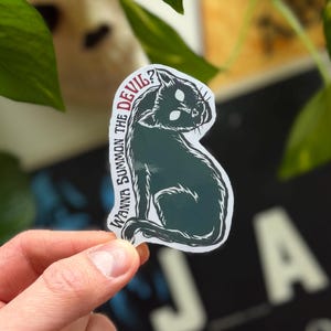 May include: A black cat sticker with the text "Wanna Summon the Devil?" in white. The cat is in a sitting position with glowing white eyes. The sticker is held in a hand against a background of green leaves and a blurred image.