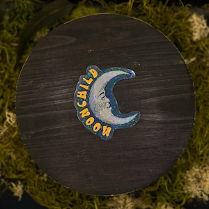 May include: A crescent moon sticker with a holographic finish and a vintage-style face illustration. The word MOONCHILD is printed in orange capital letters along the curved edge of the moon, which features a silver, glittery, star-speckled texture.