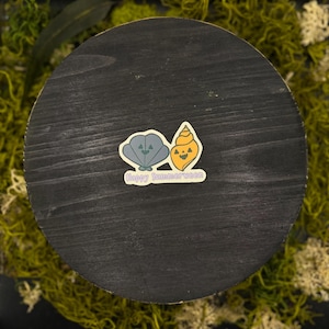 May include: A circular black wooden surface featuring a sticker with a grey clam and an orange spiral seashell, both designed with jack-o'-lantern faces. The sticker includes the text Happy Summerween in light purple lettering at the bottom.