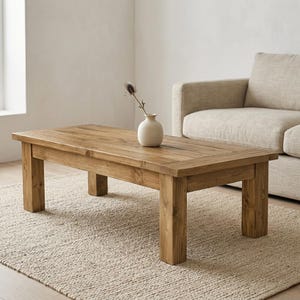May include: A rectangular, light brown wooden coffee table with a natural finish. The table has a planked top and sturdy legs. A small, white vase with a dried flower sits on the table. The table is approximately 48 inches long.