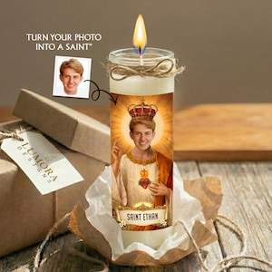 May include: A cylindrical glass candle with a lit wick, featuring a custom graphic of a person wearing a crown and religious robes with the text SAINT ETHAN. The candle is wrapped in twine and sits in a paper-lined box with a tag labelled LUMORA DESIGNS.