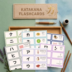 May include: A set of Katakana flashcards featuring Japanese characters, romanised text, and corresponding illustrations. The cards are organised in a wooden tray alongside a fountain pen, an inkwell, and a sprig of lavender on a blue background.
