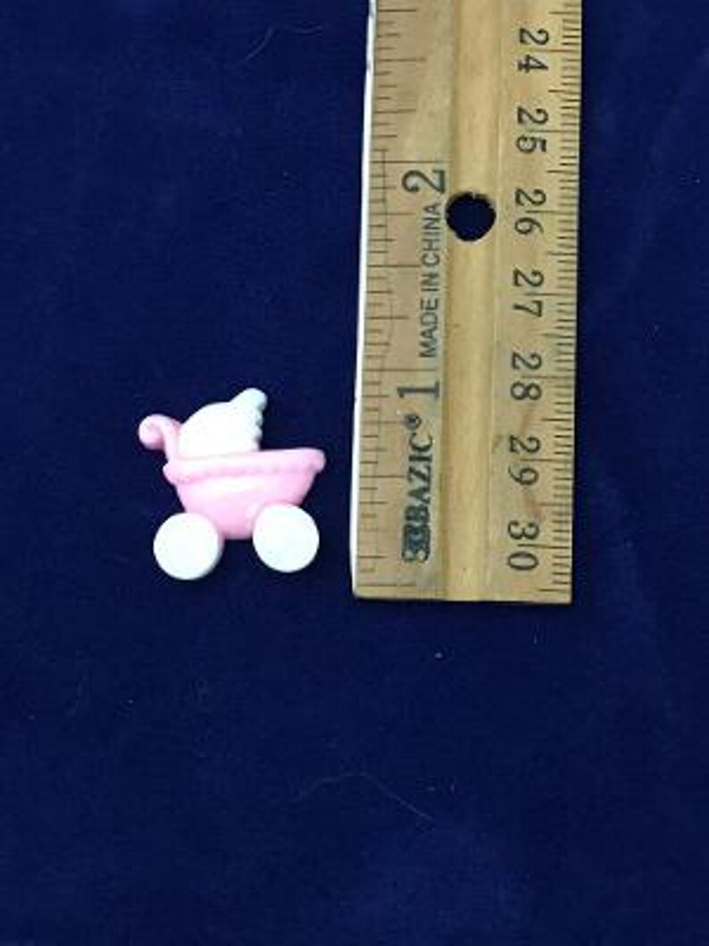 May include: A small, pink baby carriage with white wheels and a white canopy. The carriage is next to a wooden ruler with inch measurements, indicating its small size. The carriage is likely a decorative item or a cake decoration.