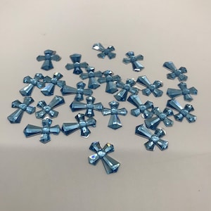 May include: A scattering of light blue, faceted, cross-shaped rhinestones. The rhinestones are scattered on a white surface.