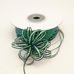 May include: A spool of green and black ribbon with a decorative bow. The ribbon has a shimmering, iridescent quality. The bow is made from the same ribbon and is in the foreground. The spool is white with a gray center.
