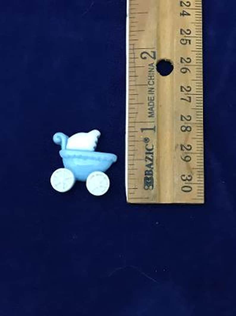 May include: A light blue miniature baby carriage with white accents and wheels. The carriage is next to a wooden ruler, showing its small size. The carriage is approximately 1 inch in length.