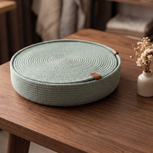 Op de afbeelding: Een ronde, lichtgroene geweven touwmand met een spiraalvormig ontwerp. De mand heeft een bruin leren lipje aan de zijkant. Hij staat op een donkerbruine houten tafel. Een kleine witte vaas met gedroogde bloemen staat op de achtergrond.
