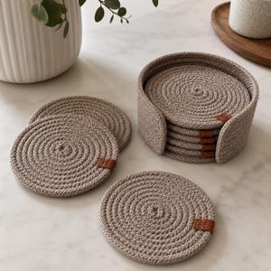 May include: A set of round, beige rope coasters with a spiral pattern. Each coaster features a small, brown leather-like tab stitched to the edge. The coasters are stacked inside a matching cylindrical rope holder, with two additional coasters placed flat.