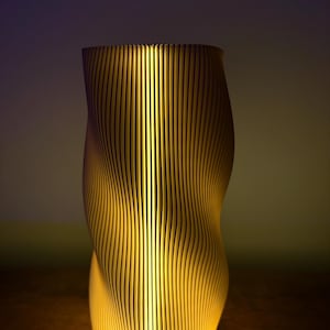 Può includere: Una lampada moderna e illuminata con un design unico. La lampada è realizzata con doghe di legno verticali che si incurvano e si torcono, creando un motivo ondulato. L'interno si illumina con una luce calda e gialla, proiettando ombre e mettendo in risalto la trama del legno.