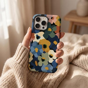 Puede incluir: Una funda de teléfono con un vibrante estampado floral en tonos azules, amarillos, rosas y verdes sobre un fondo azul oscuro. La funda tiene un acabado brillante y está sujeta con una mano.