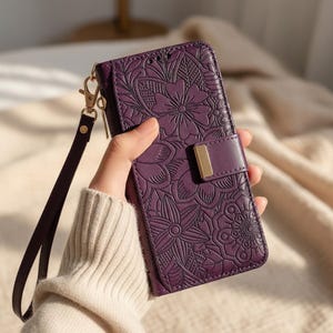 Puede incluir: Una funda de teléfono morada con un diseño floral en relieve. La funda tiene una correa para la muñeca y un cierre dorado. La funda está hecha de un material similar al cuero y tiene un cierre magnético.