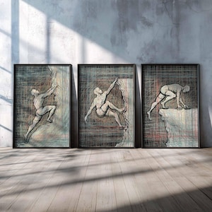 May include: Set of three framed art prints depicting a climber in various stages of ascent. The artwork features a sketch-like style with a muted color palette of gray, blue, and red. The frames are black, and the prints are displayed against a gray wall and wooden floor.