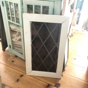 May include: A white-framed cabinet door with a diamond-patterned window pane. The door is leaning against a wooden floor. A light blue cabinet with glass doors is in the background.