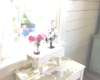 Op de afbeelding: Een witte houten tafel met een verweerde afwerking staat naast een raam. Twee kleine witte houten krukjes zijn op de tafel gestapeld. Een vaas met roze rozen staat op het bovenste krukje. Een zwarte decoratieve metalen standaard met een kristallen kroonluchter staat op het onderste krukje.