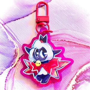 May include: A red keychain featuring a cartoon lamb character with a white fluffy head, black body, and a red cape. The lamb has a gold bell around its neck and is set against a pink starburst shape.