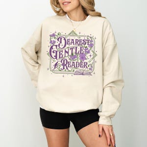 May include: A beige sweatshirt with the text "Dearest Gentle Reader" in purple script and floral accents. The design includes an open book and geometric shapes. The sweatshirt is a casual style.