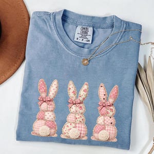 Pode incluir: Uma camiseta Comfort Colors azul claro com três coelhos da Páscoa aplicados. Cada coelho é feito de tecido rosa estampado e tem um laço rosa e uma rosa branca na base. Um colar de ouro está drapeado sobre a camisa.
