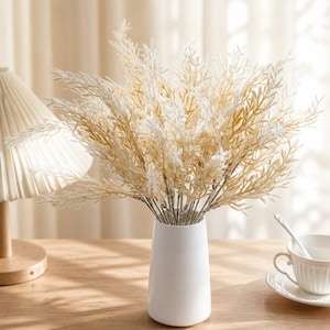 May include: A white ceramic vase containing a bouquet of artificial plant stems with delicate, feathery foliage in shades of cream and light tan. The stems are arranged in a dense, upright cluster, showcasing fine, branching textures throughout.