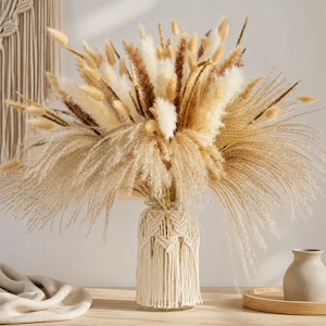 Peut inclure: Un vase en verre enveloppé d'une corde en macramé crème avec un motif à chevrons et des franges, rempli d'un grand arrangement d'herbes de la pampa séchées, de queues de lapin blanches duveteuses et de tiges brunes dans des tons beige et ocre.
