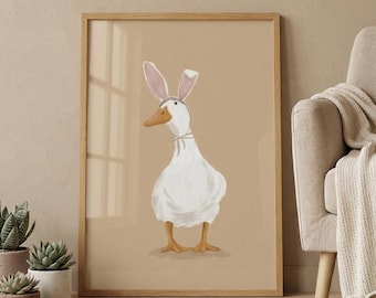 May include: A framed print featuring a whimsical illustration of a white goose wearing bunny ears. The goose has an orange beak and feet, set against a tan background. The artwork is displayed in a light brown wooden frame, with a neutral-toned interior setting.