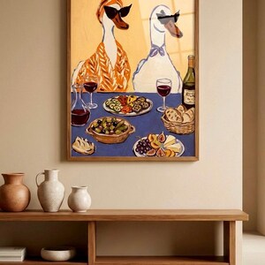 May include: A framed art print depicts two anthropomorphic geese wearing sunglasses, seated at a table laden with food and wine. The artwork features a blue tablecloth, a bottle of wine, and various dishes. The overall color palette is warm, with a wooden frame.