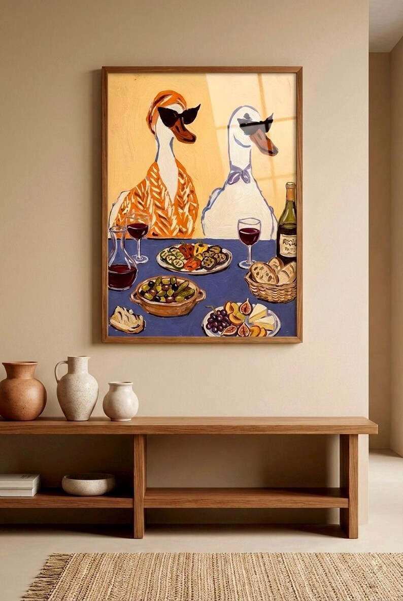 May include: A framed art print depicts two anthropomorphic geese wearing sunglasses, seated at a table laden with food and wine. The artwork features a blue tablecloth, a bottle of wine, and various dishes. The overall color palette is warm, with a wooden frame.