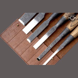 May include: A set of woodworking chisels with wooden handles and metal blades, displayed in a brown leather tool roll. The chisels have a rustic, hammered metal finish. The tool roll is open, revealing the tools.