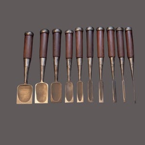 May include: A collection of ten woodworking chisels. Each chisel has a dark wooden handle with a silver-colored metal ferrule and blade. The blades vary in shape and width, with some having flat ends and others pointed.