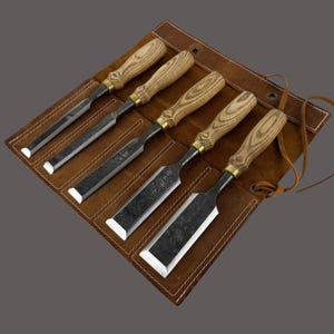 Puede incluir: Un juego de cinco formones para madera con mangos de madera clara y casquillos de latón, dispuestos en un estuche de cuero marrón. Cada formón presenta una hoja de metal oscuro y texturizado con un borde afilado y biselado en ranuras de cuero.